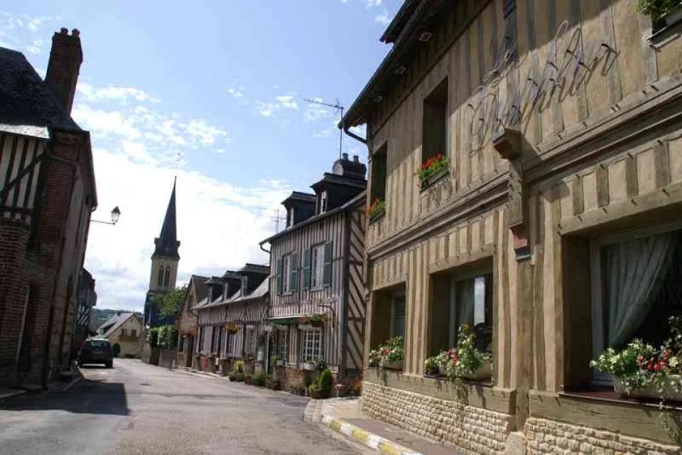 rue-colombages-le-breuil-en-auge-calvados-ot-terre-d-auge Rue typique du Breuil-en-Auge avec maisons à colombages et clocher à l’arrière-plan.